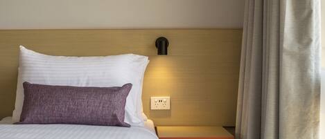 Una cama bien hecha con almohadas blancas y moradas, una lámpara en la pared y una mesita de noche de madera.