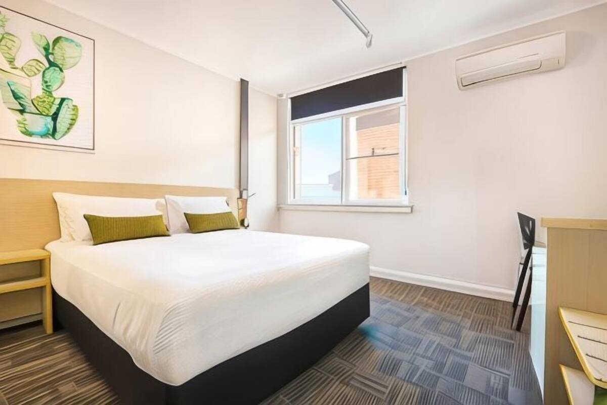 A hotel room with a bed, a desk, a chair, a window, and an air conditioning unit.