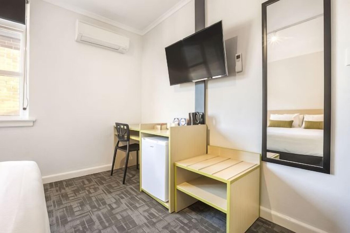 A compact hotel room with a bed, a desk with a chair, a small refrigerator, a television mounted on the wall, and a large mirror reflecting the bed.