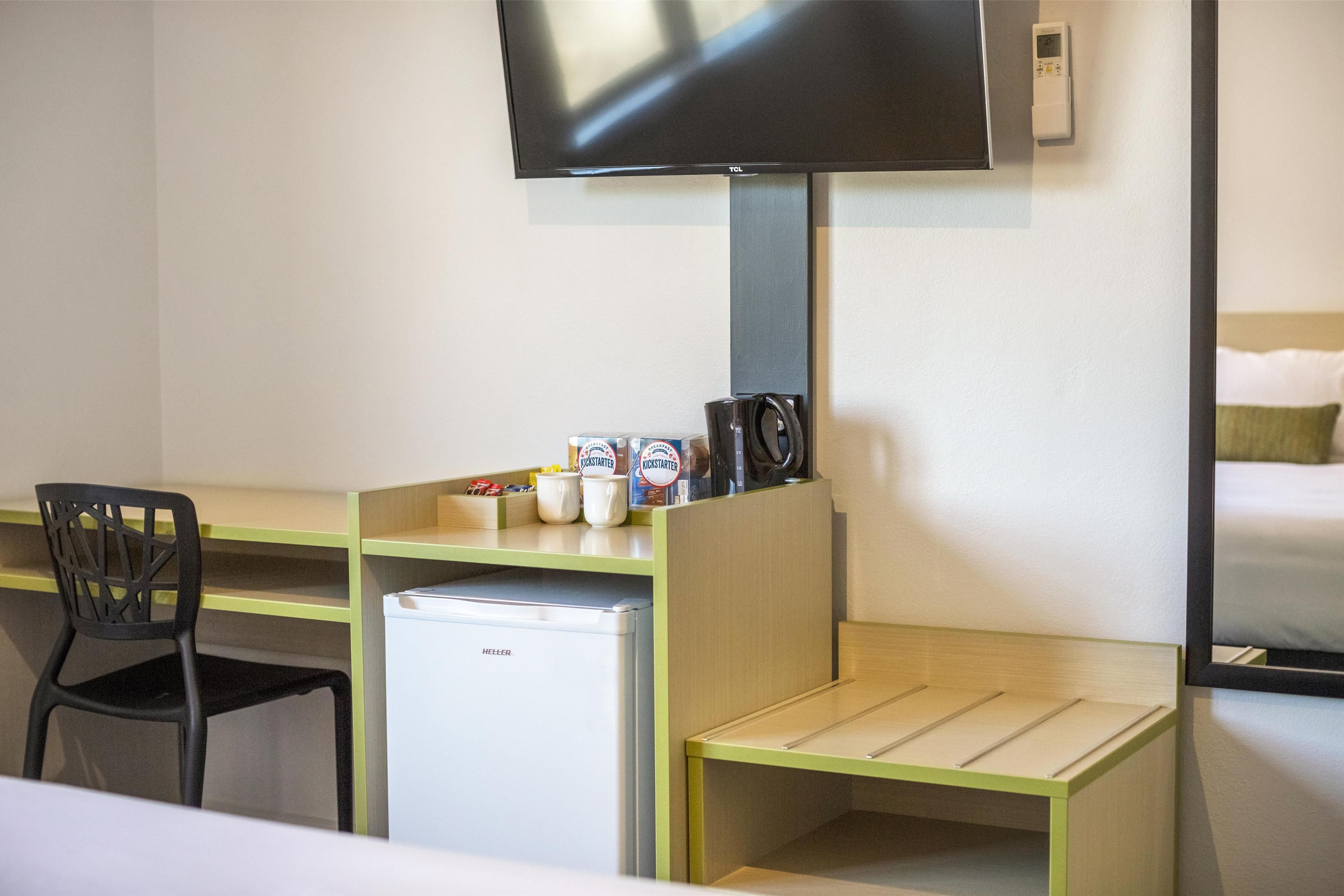 A compact hotel room with a desk, chair, small refrigerator, and a television mounted on the wall.