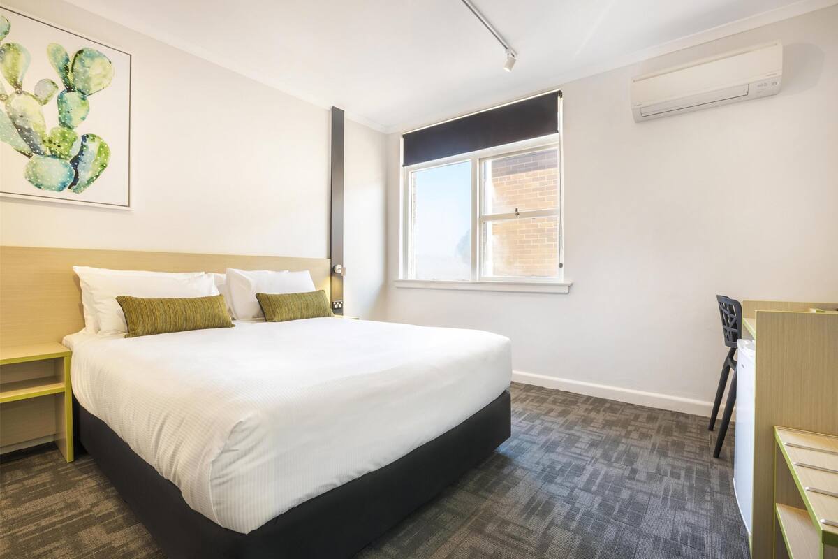 A modern hotel room with a large bed, a desk, and a window with a view of a brick building.