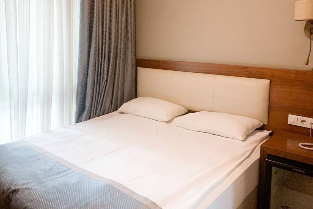 Ein Hotelzimmer mit Bett, Holz-Kopfstütze, Nachttisch mit Lampe und einem Fenster mit Vorhängen.