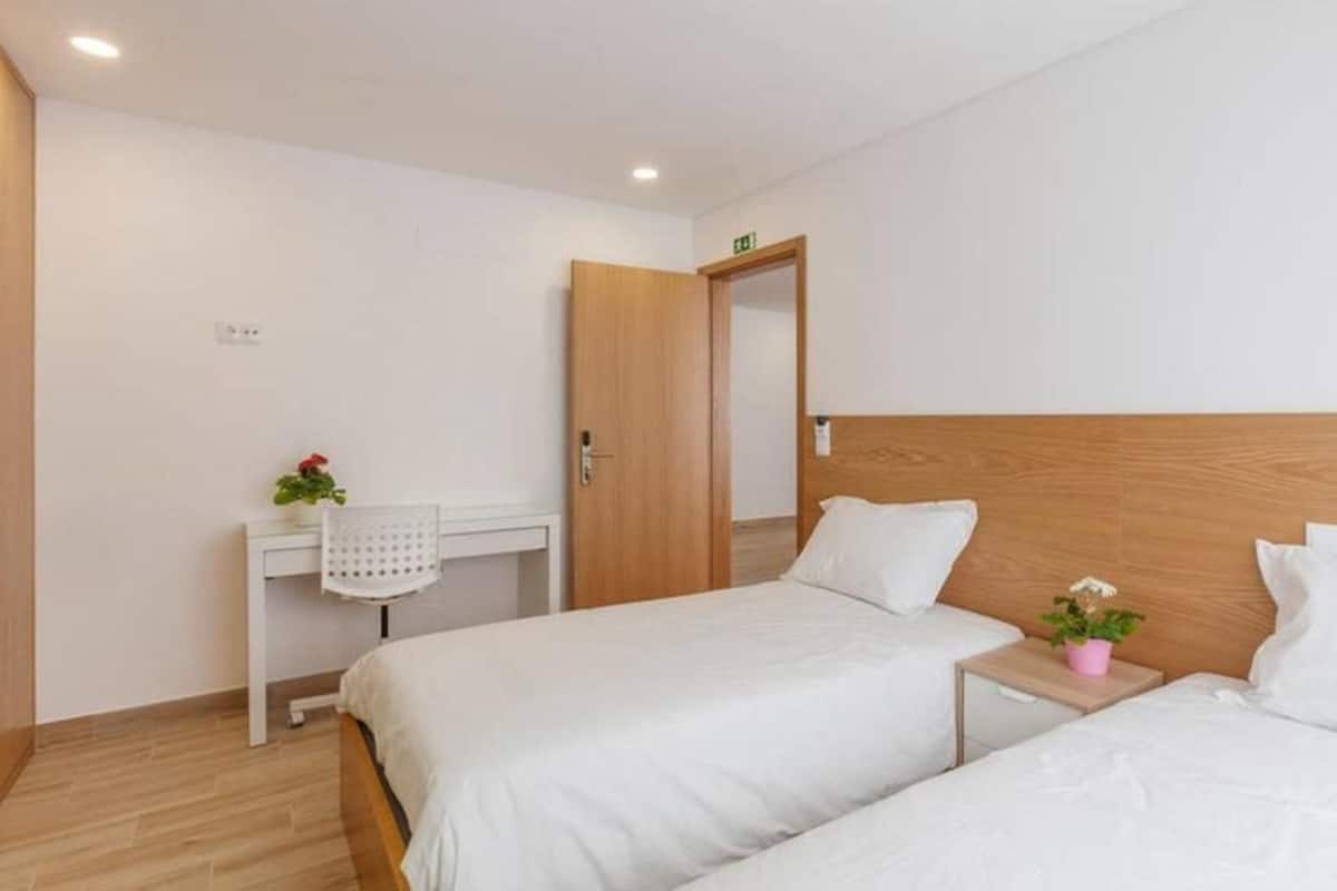 Habitación de hotel con dos camas, cabecero de madera, un escritorio pequeño con silla y una planta en maceta.