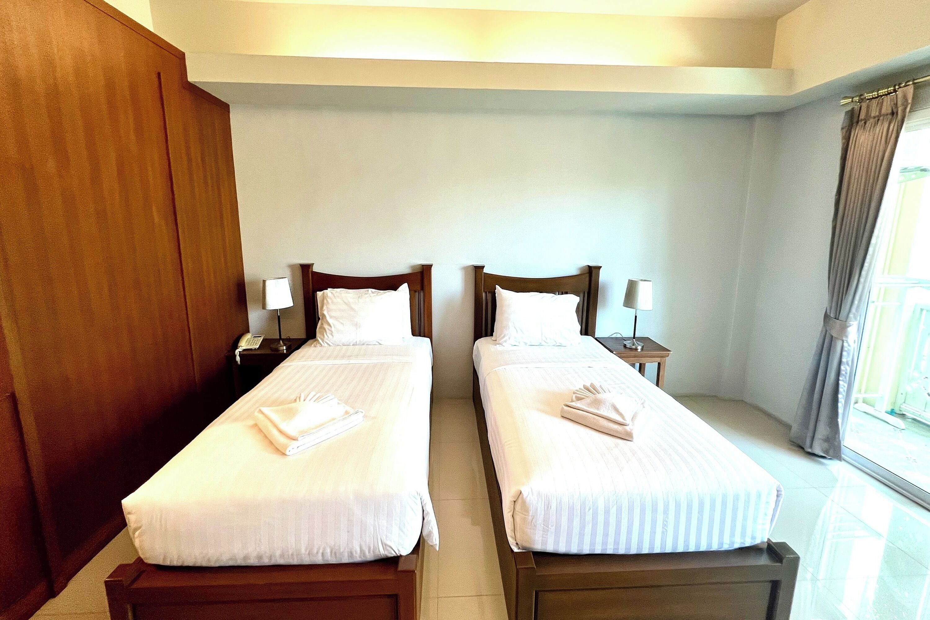 Habitación de hotel con dos camas individuales, cada una con una almohada y una toalla doblada. Se observan mesitas de noche con lámparas y un teléfono.