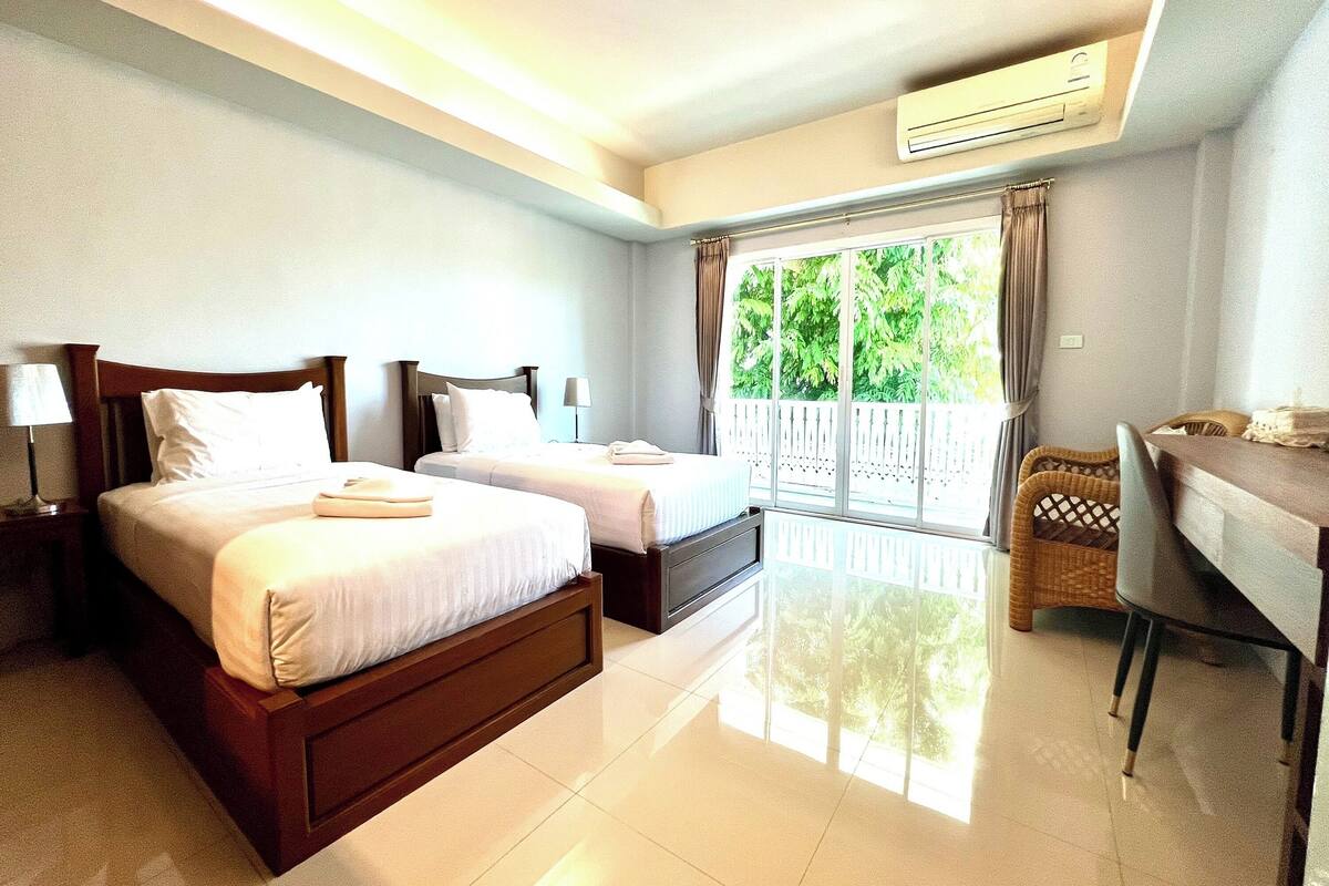 A hotel room with two beds, a desk, a chair, and a balcony with a view of greenery.