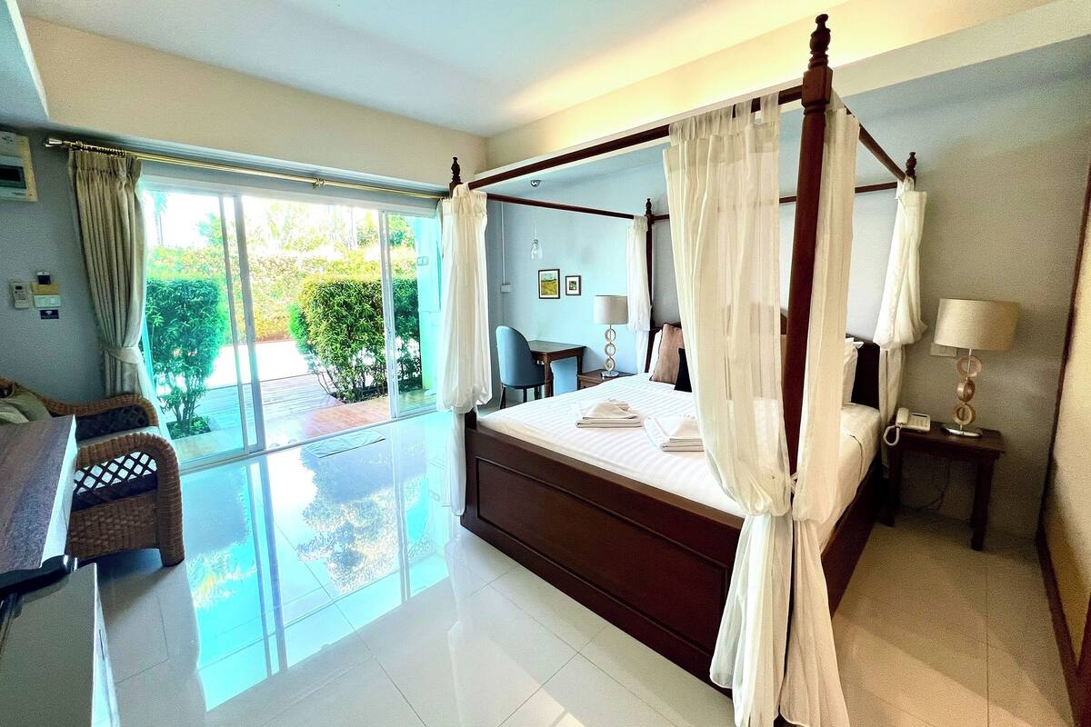 A four-poster bed with white linens, a bedside table with a lamp, and a view of greenery through large sliding glass doors.