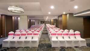 A hotel conference room with rows of white chairs with pink sashes, a patterned carpet, and a modern ceiling design.