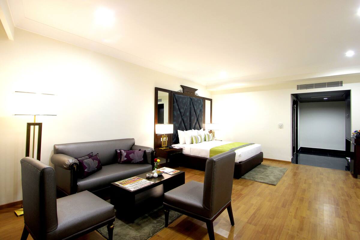 Habitación de hotel con sofá, sillón, una mesita y cama con colcha verde.