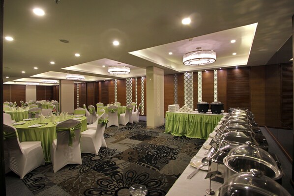 Un salón de banquetes con mesas redondas cubiertas con manteles verdes, sillas blancas con cintas verdes y una larga barra de buffet con estaciones de servicio de alimentos.
