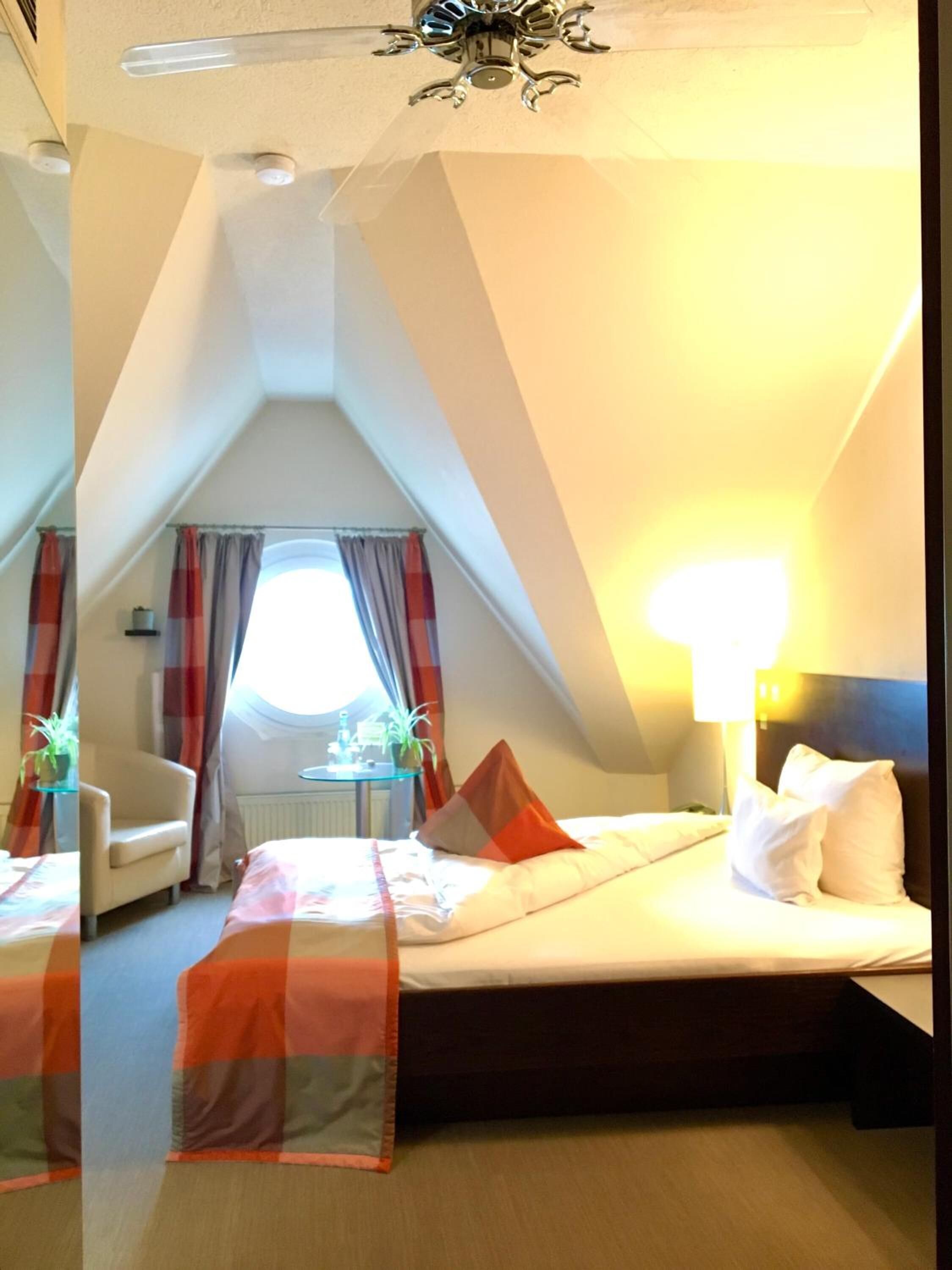 A hotel room with a bed, a chair, a table with a plant, and a window with curtains.