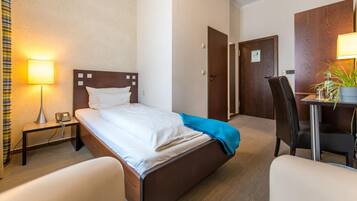 Habitación de hotel con cama, mesita de noche con lámpara, escritorio y silla, y una puerta.