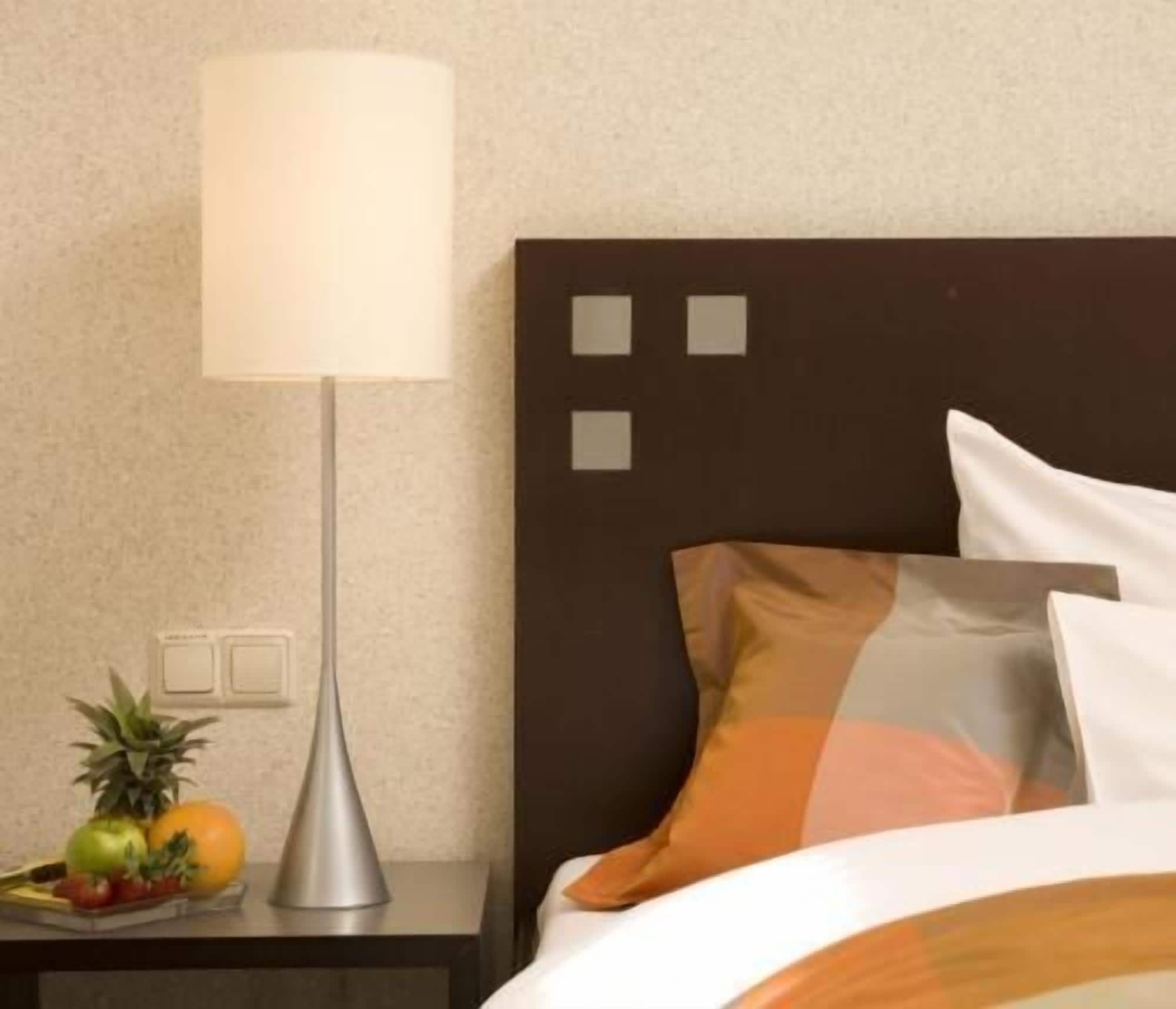 Une chambre d’hôtel bien rangée, avec un lit, une table de chevet dotée d’une lampe et un panier de fruits.