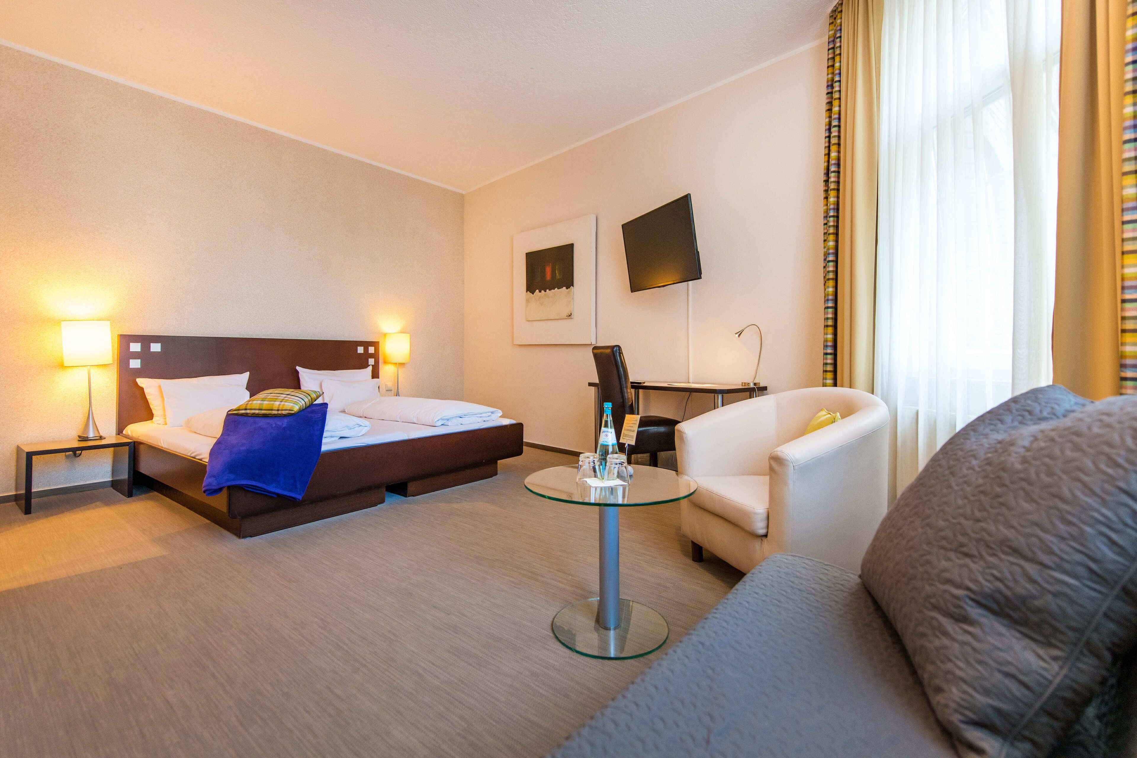 A hotel room with a bed, a sofa, a glass coffee table, and a desk with a chair.