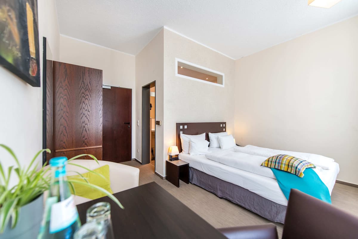 A hotel room with a bed, bedside table, lamp, and a small table with a plant and glasses.