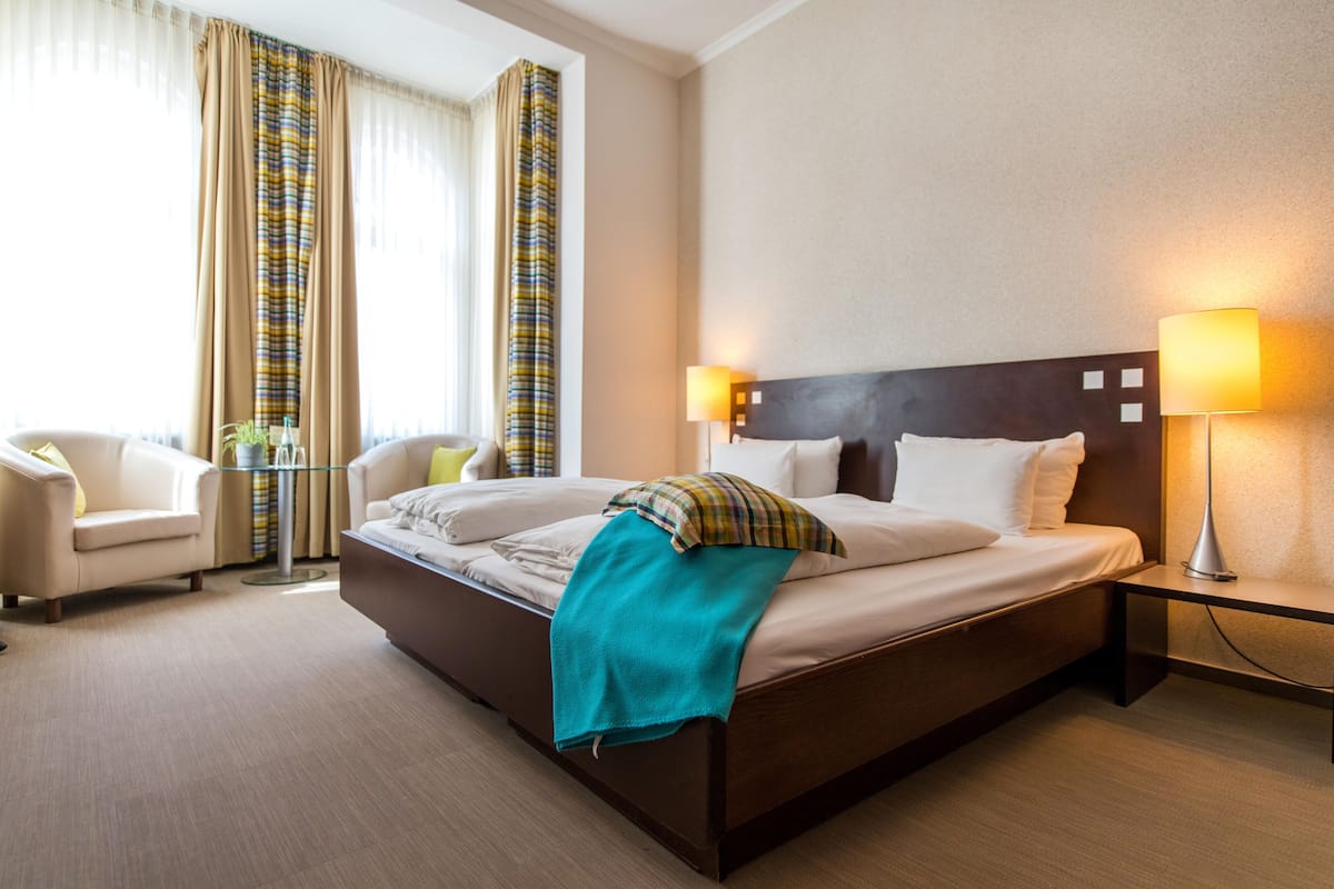 A hotel room with a large bed, two bedside tables with lamps, a chair, and a window with curtains.