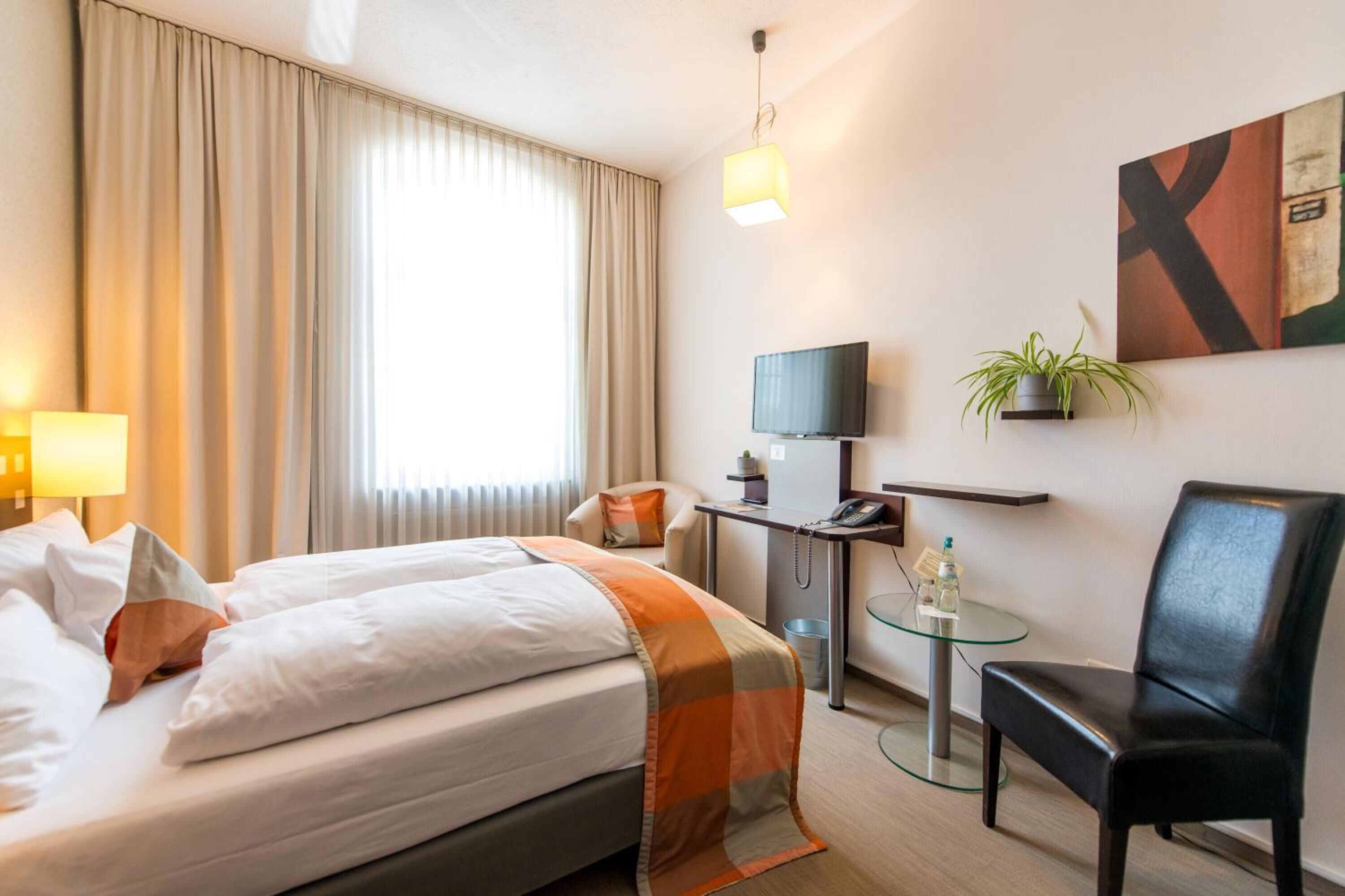 Une chambre d’hôtel comprenant un lit, un bureau avec un ordinateur, une chaise, une petite table, une plante et un téléviseur.