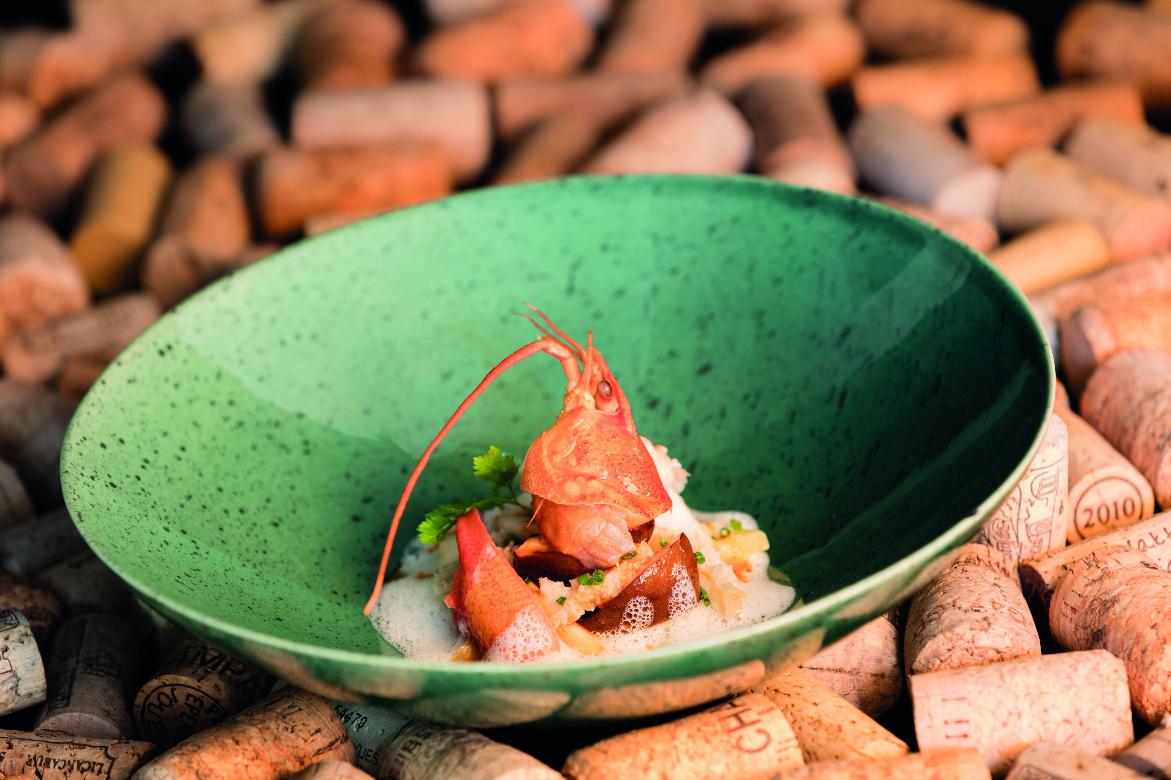 A green bowl with a lobster on a bed of what appears to be rice and vegetables, surrounded by wine corks.
