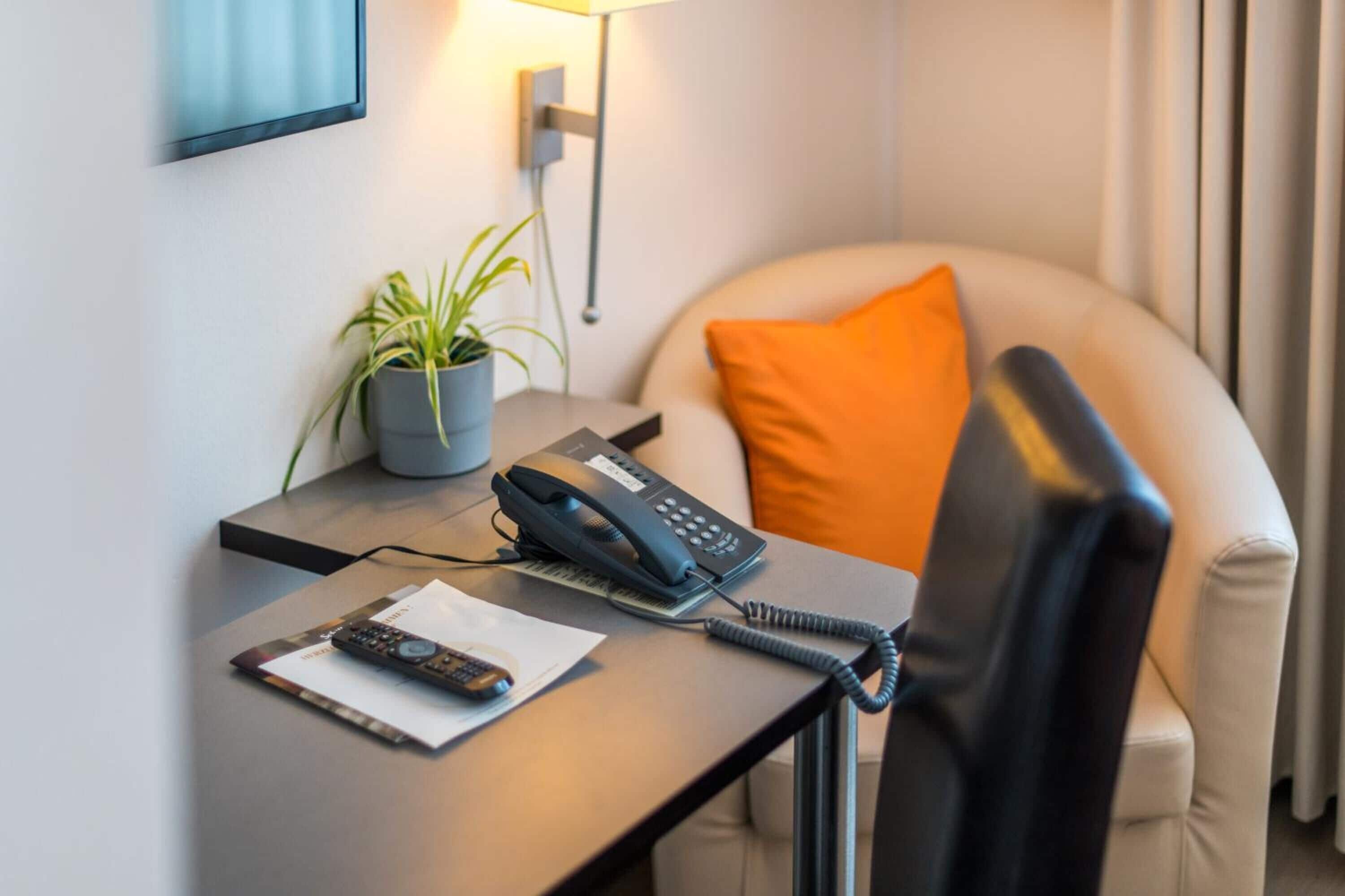 Habitación de hotel con escritorio, teléfono, control remoto, planta y cojín.
