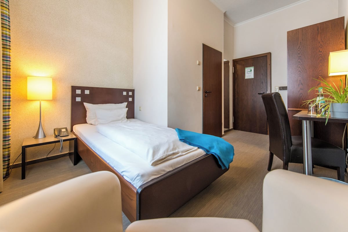 A hotel room with a bed, bedside table with a lamp, a desk with a chair, and a door.