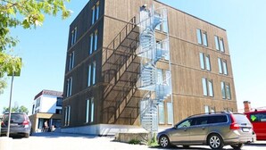 Un edificio de varios pisos con una escalera de caracol en el lateral, coches aparcados y cielo despejado.