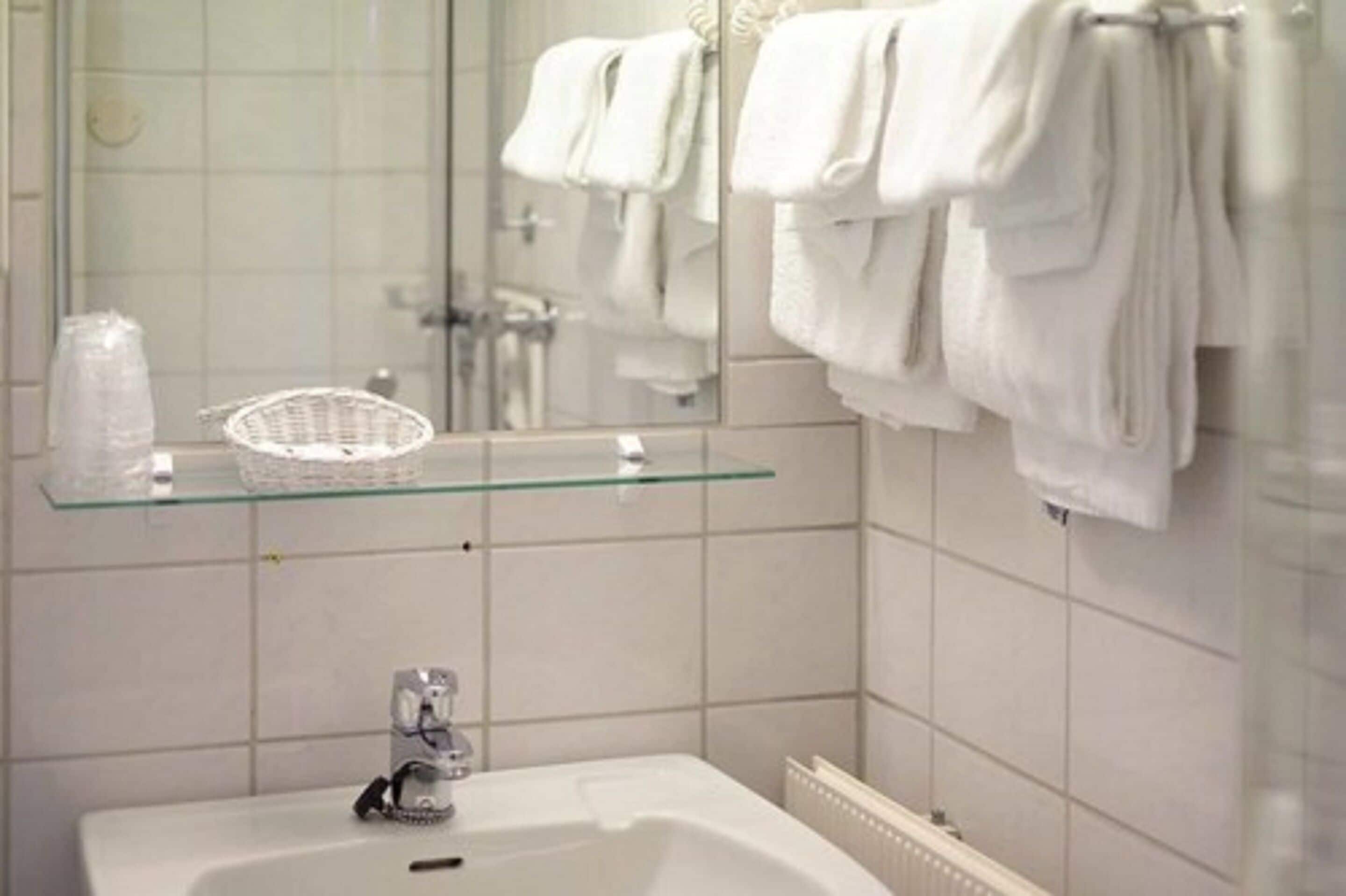 Egy fürdőszoba, melyben egy üvegpolcon kosár és egy üveg található, emellett van egy fehér mosdókagyló és egy törölközőtartó, melyen több törölköző is van.