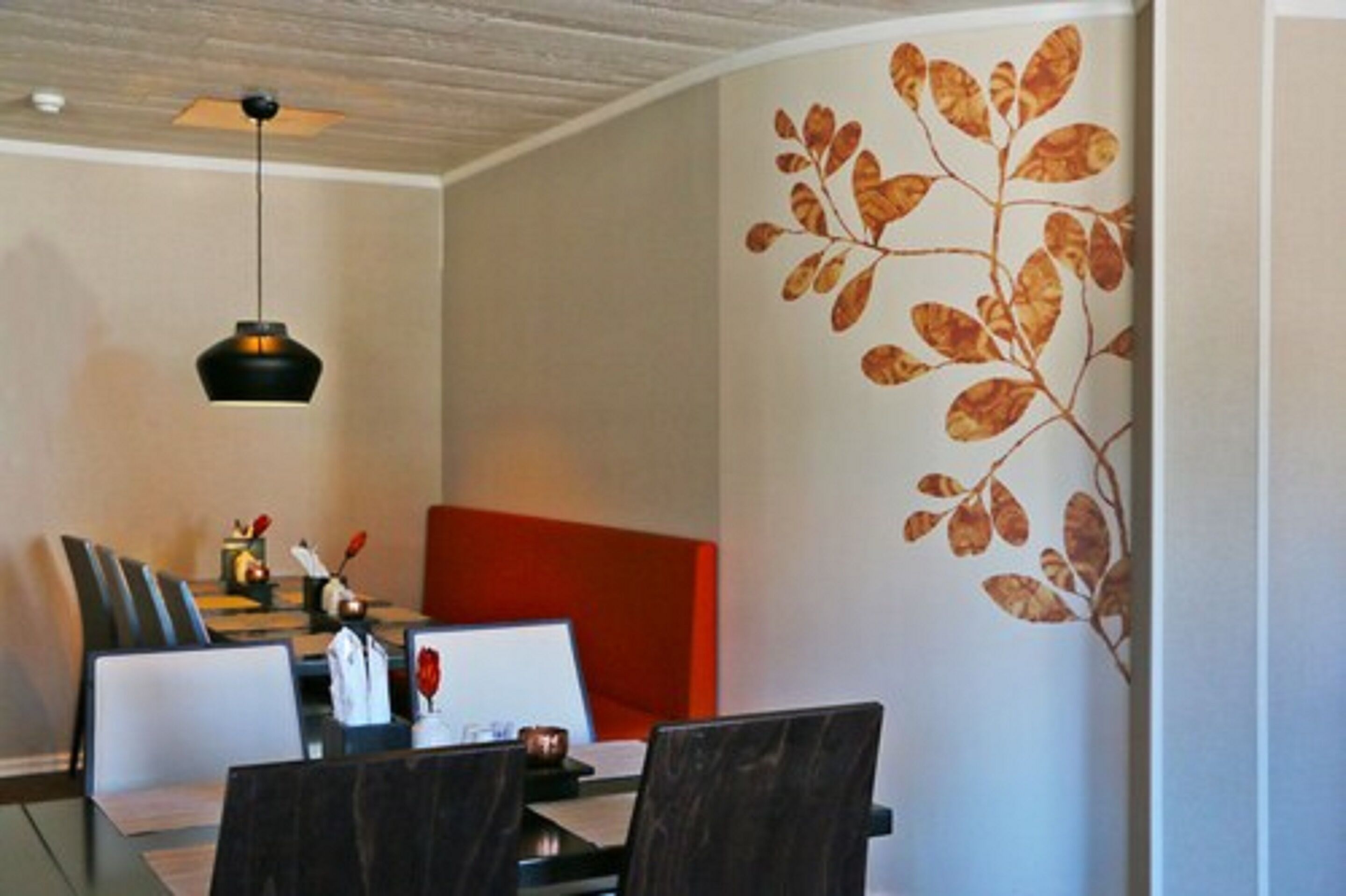 A dining area with a table, chairs, a red cushioned bench, a pendant light, and a wall with a leaf pattern.