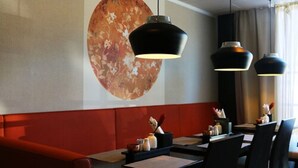 Interior de restaurante con un gran cuadro abstracto en la pared, iluminación colgante negra y un banco con cojines rojos.