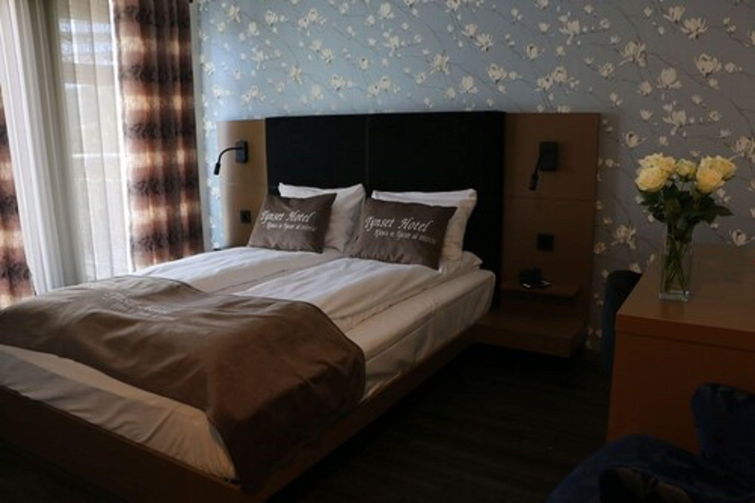 A hotel room with a large bed, two pillows with text, a nightstand, a vase with flowers, and a wall with a floral pattern.