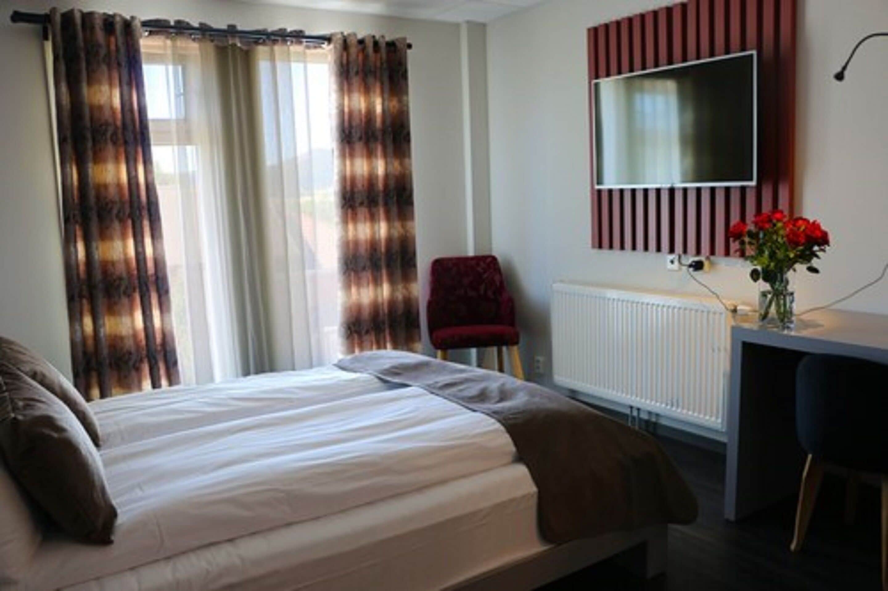 A bedroom with a bed, a red chair, a desk with a TV, and a radiator.