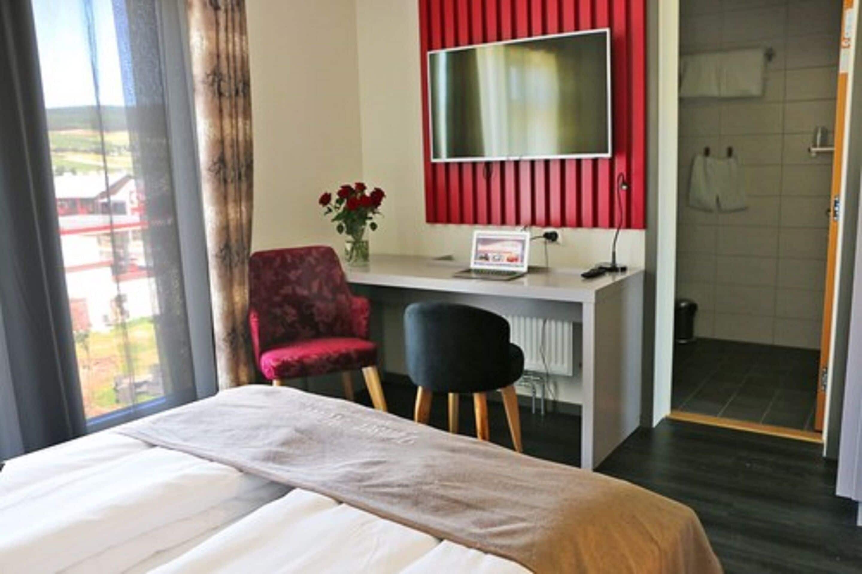 A hotel room with a bed, a desk with a computer, a TV, a chair, and a view of the outside.