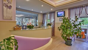 A reception area with a curved counter, potted plants, and a television on the wall. - Hotel Europa (Sorico)