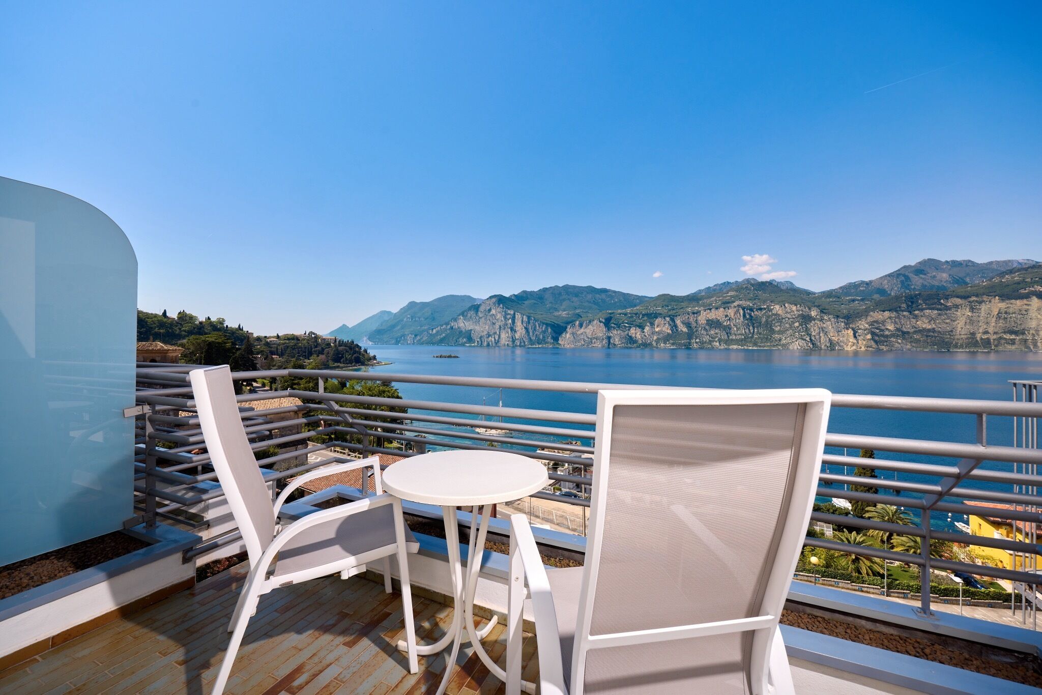 Ein Balkon mit weißen Stühlen und einem Tisch mit Blick auf ein Gewässer und Berge.