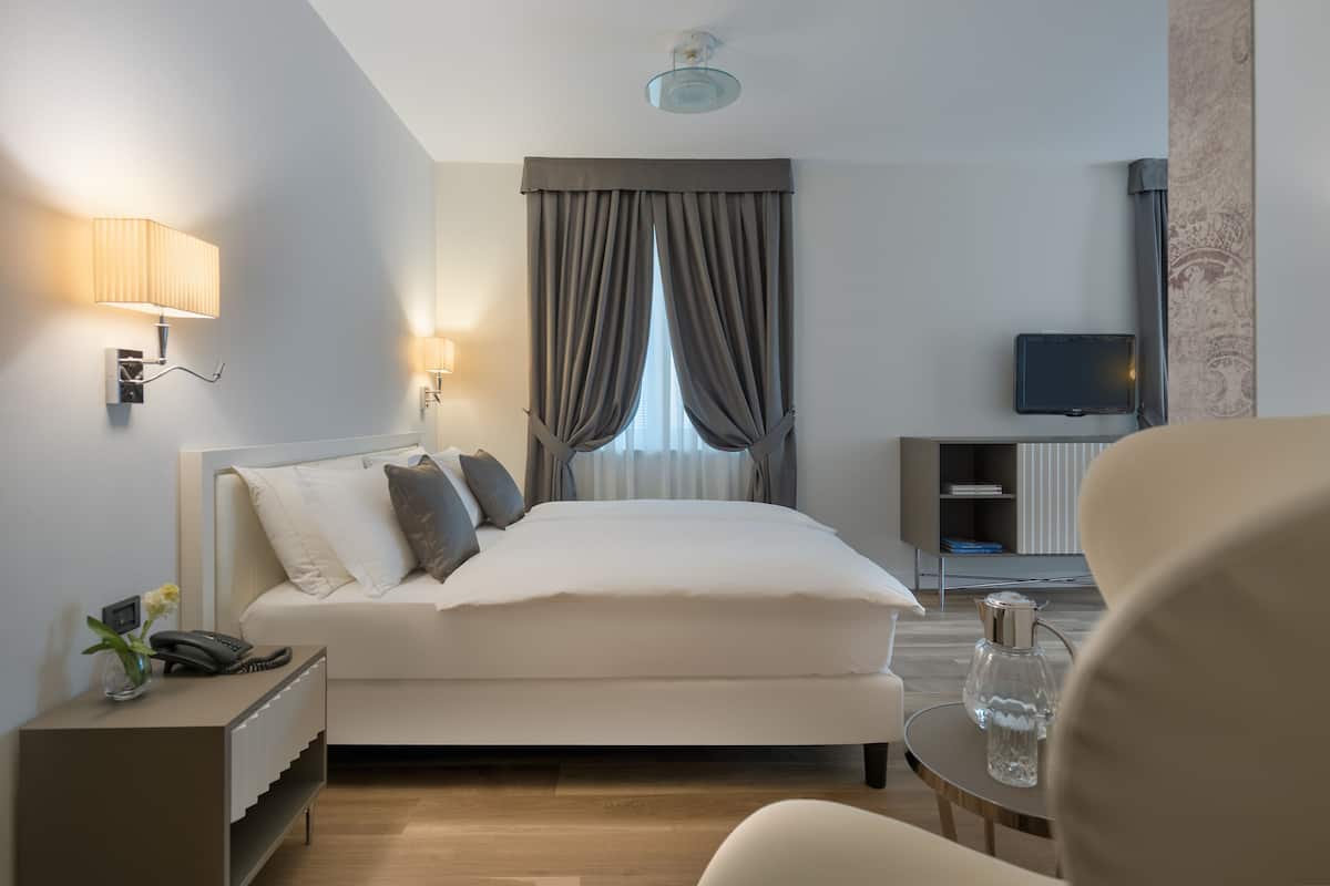 Habitación de hotel moderna con cama, lámpara de noche, una mesita con un florero, televisor sobre un mueble y vista al exterior a través de la ventana.