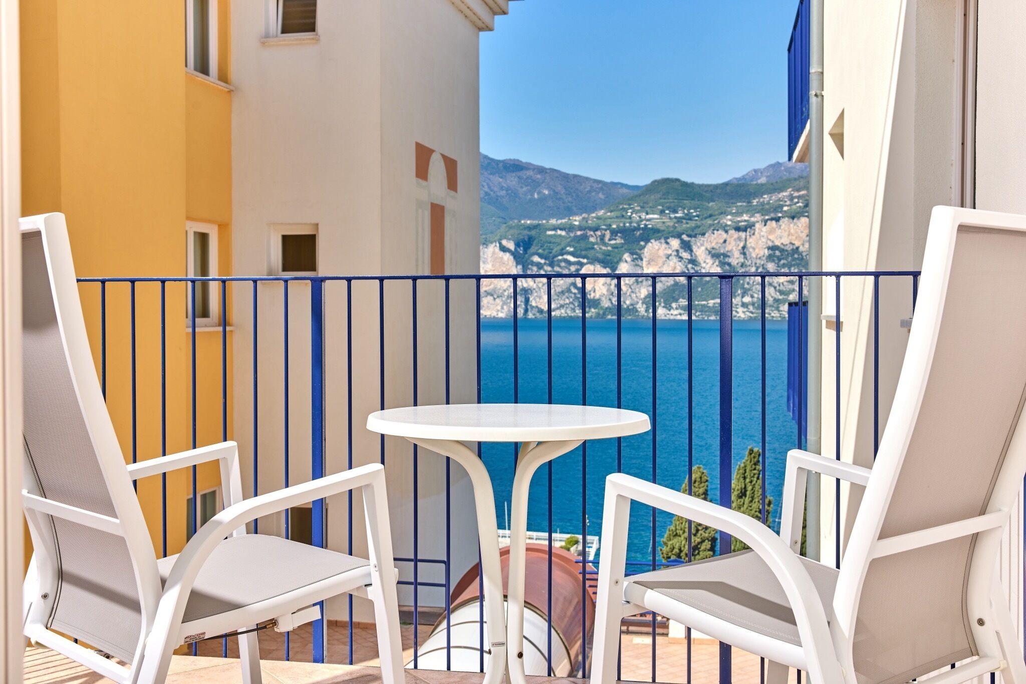 A balcony with white chairs and a small table, overlooking a coastal view with cliffs and the sea.
