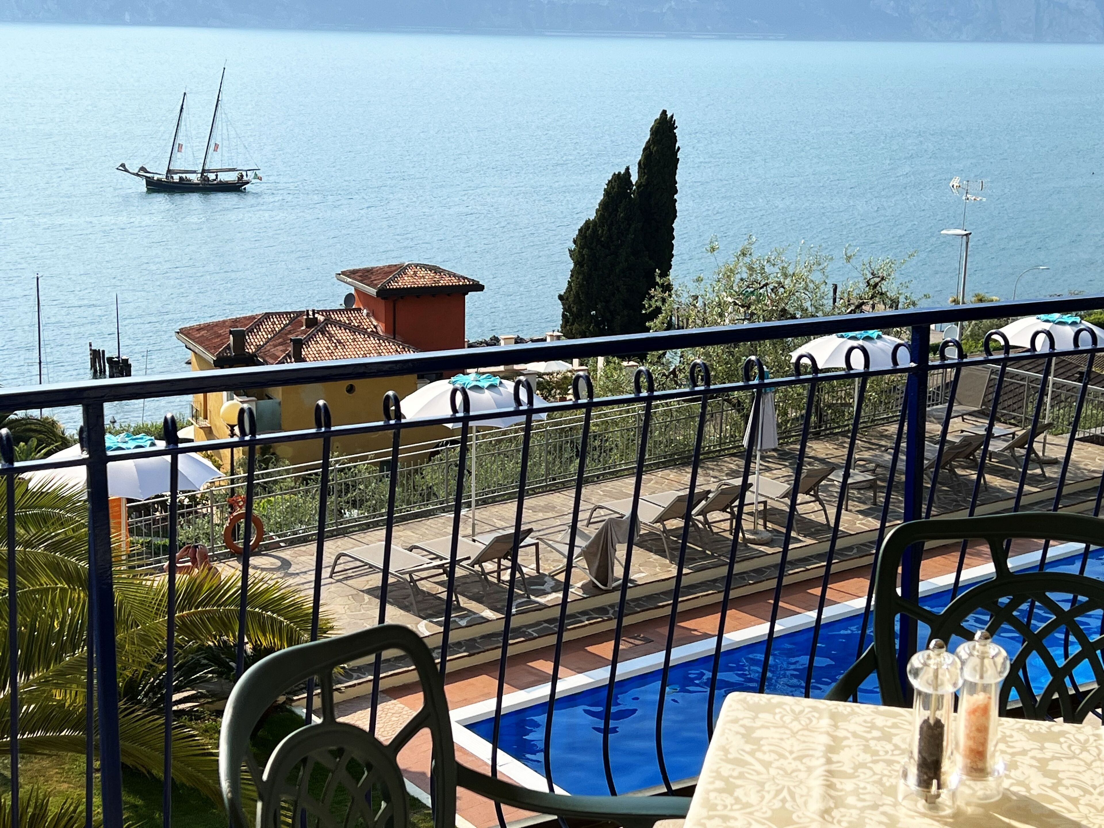 A poolside view with a clear view of the sea, a sailboat, and a building with a tiled roof.
