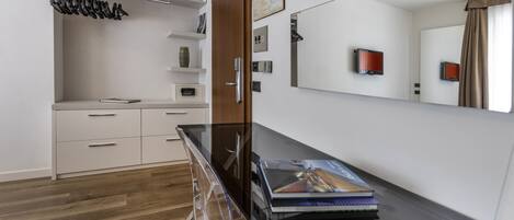 A modern hotel room with a wooden floor, a glass desk, a large mirror, a wardrobe with hanging clothes, and a small shelf with a vase.