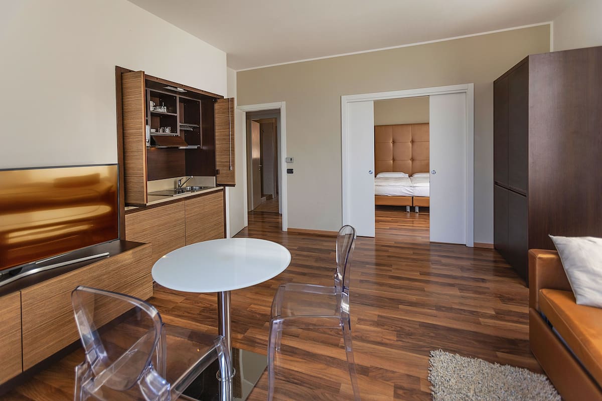 Habitación de hotel moderna con suelo de madera, una mesa redonda de comedor, sillas traslúcidas, un sofá, una cama con cabecero y una zona de cocina integrada.