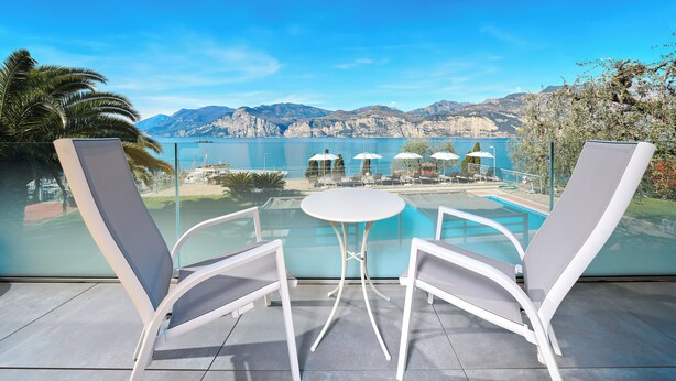 A balcony with two white lounge chairs and a small table, overlooking a pool and mountains. - Hotel Capri (Malcesine)