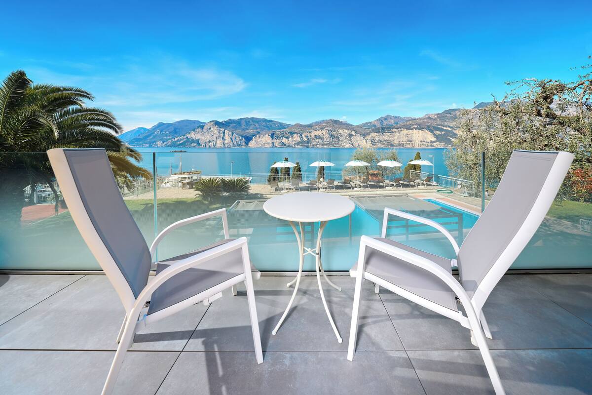Un balcón con dos sillones blancos y una mesita pequeña, con vista a una piscina y montañas.