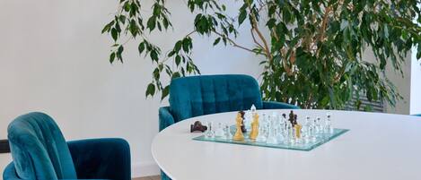 Uma mesa redonda com um tabuleiro de xadrez no centro, rodeada por cadeiras azul-turquesa, e uma planta verde grande ao fundo.
