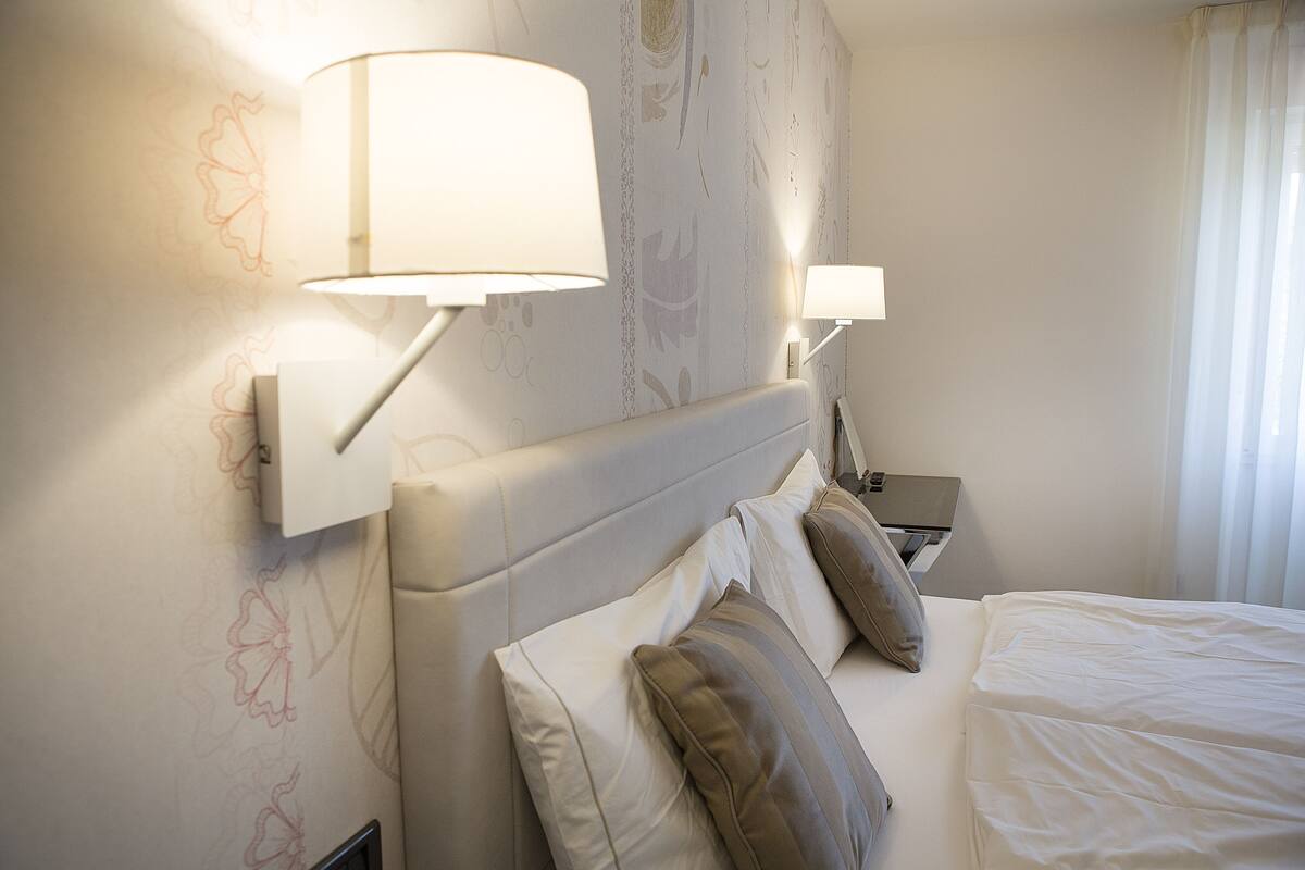 Una habitación de hotel con una cama, dos lámparas de noche, una mesita de noche y una pared con papel tapiz floral.