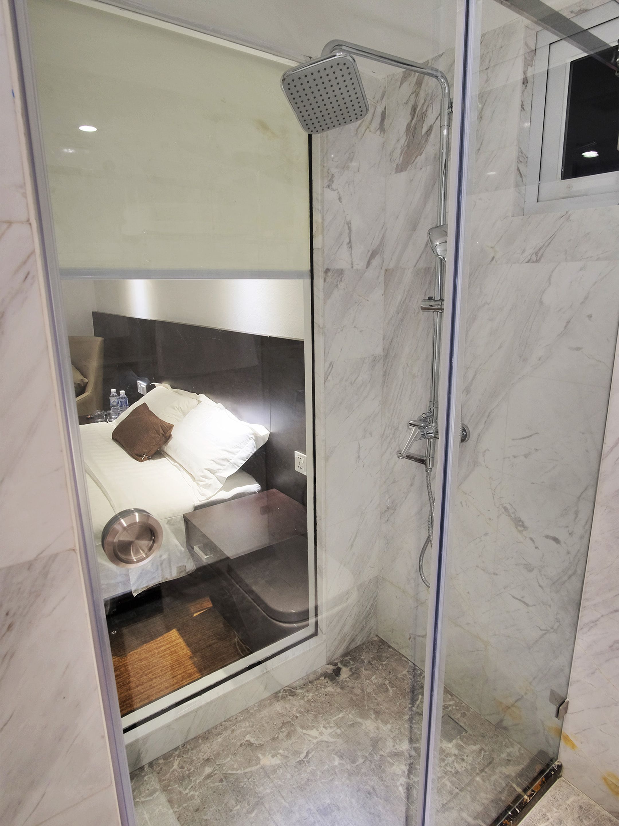 A modern bathroom with a glass shower enclosure, a marble-tiled floor, and a visible bed with a pillow and a metal bowl.