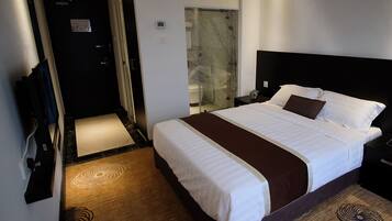 A hotel room with a large bed, a bedside table, a nightstand, a bathroom with a shower, and a door leading to another room.