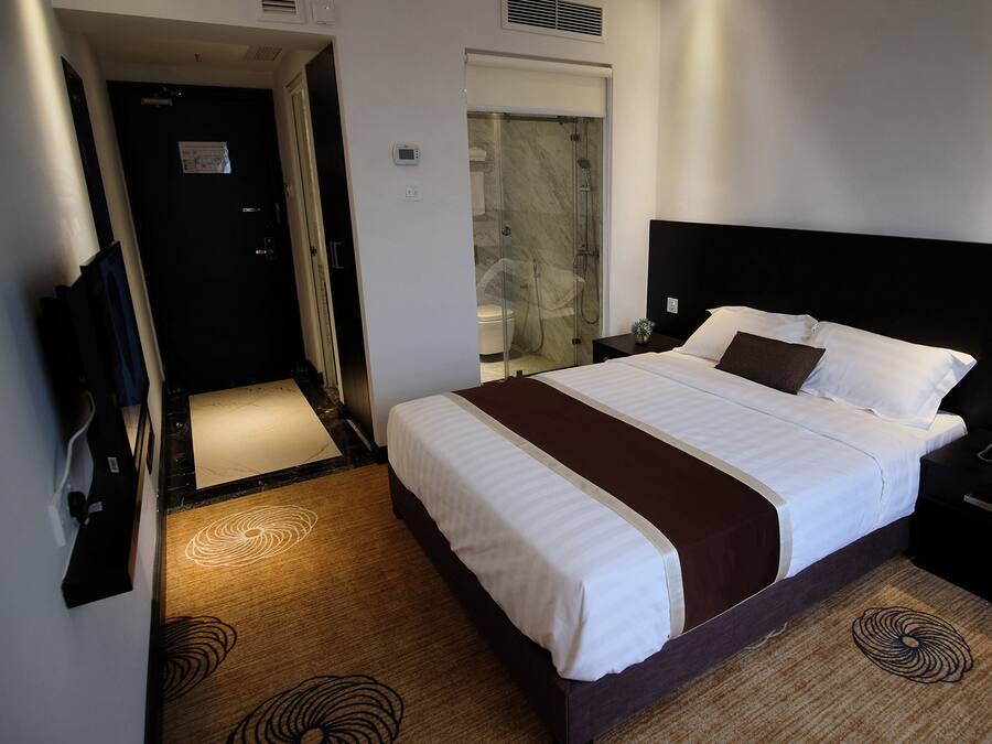 A hotel room with a large bed, a bedside table, a nightstand, a bathroom with a shower, and a door leading to another room.