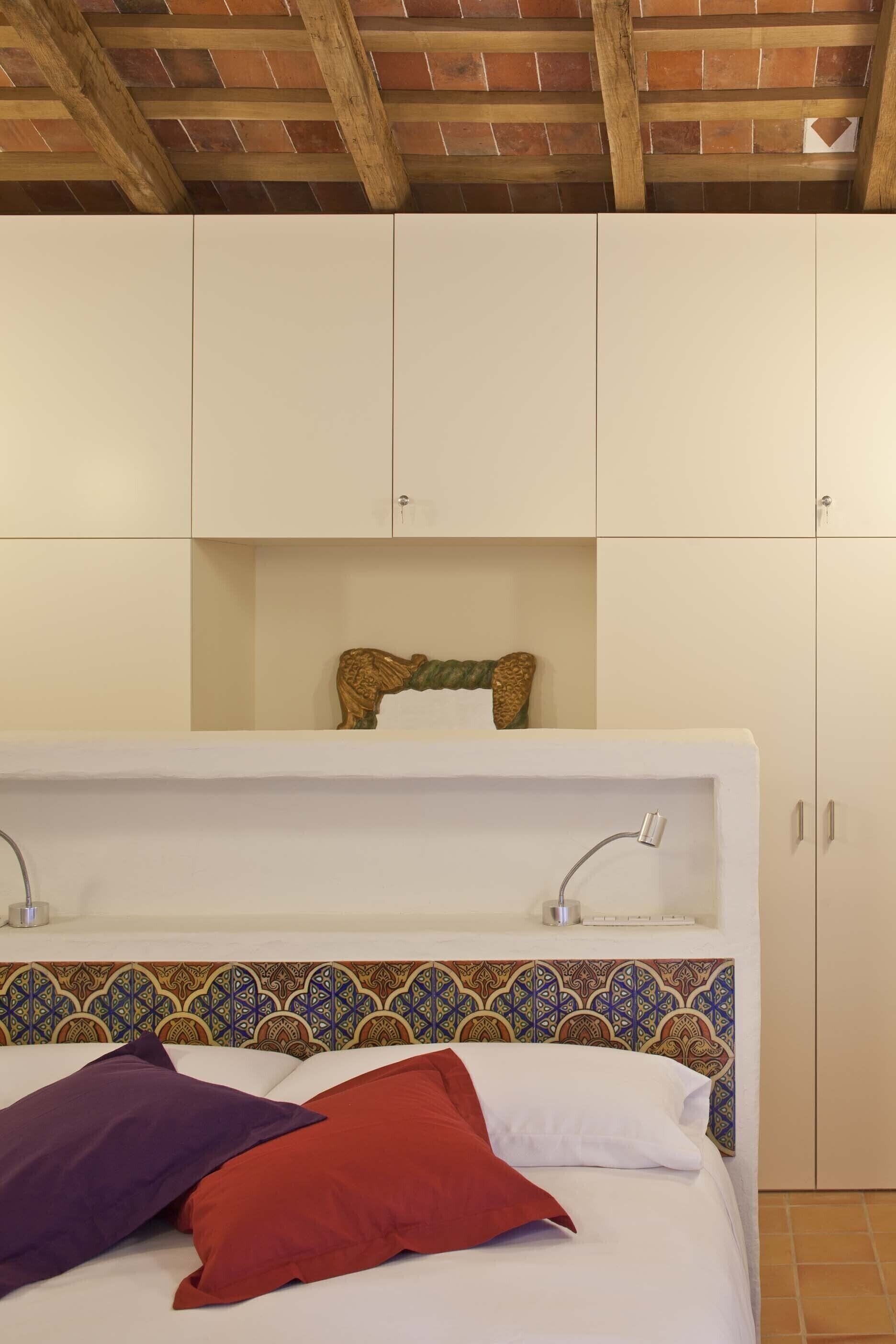 A bedroom with a bed, a headboard with a patterned design, a shelf above the bed, and a wardrobe with two doors.