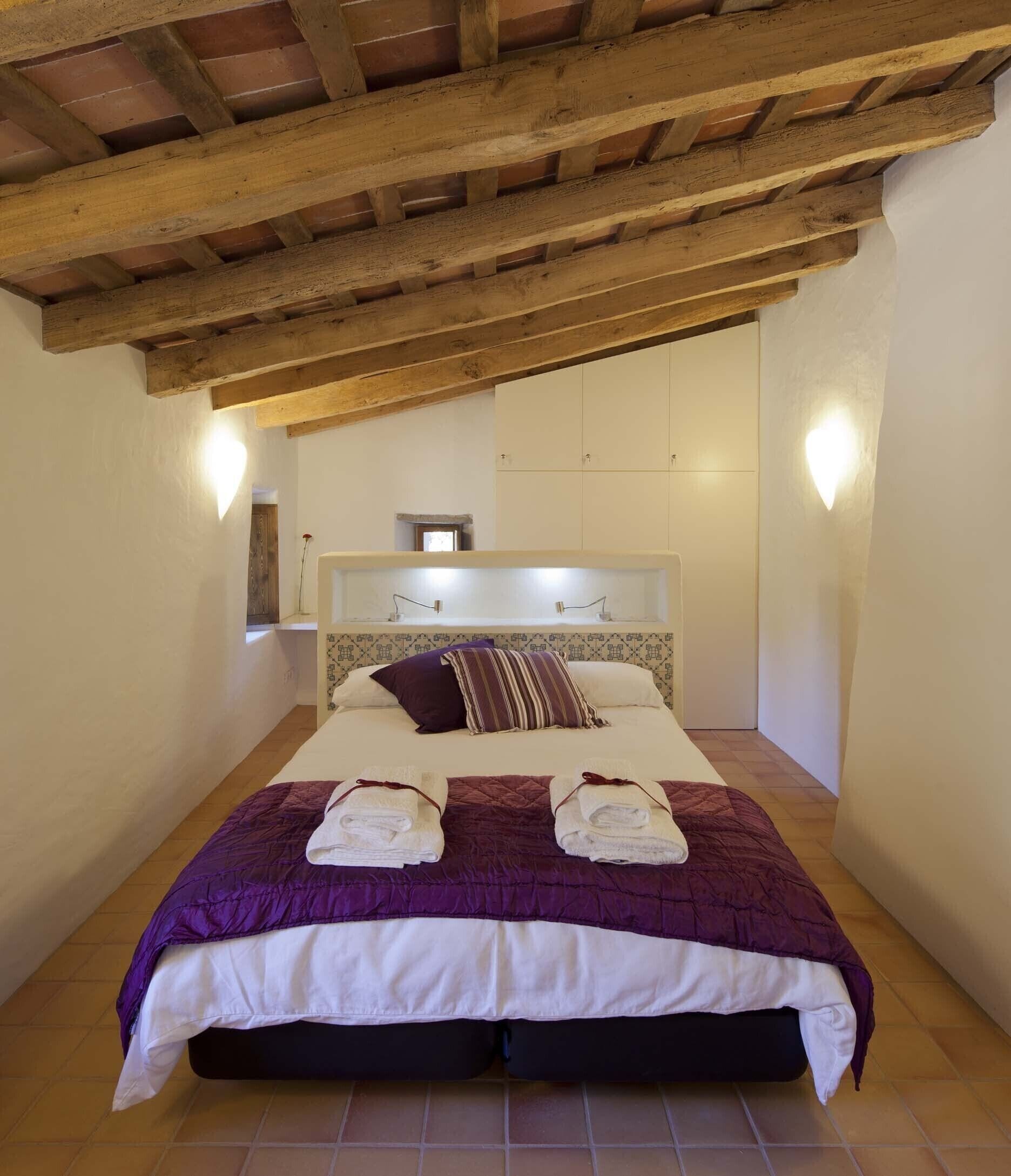 Une chambre avec un plafond en bois, un lit recouvert d’une courtepointe violette et blanche, deux serviettes sur le lit et une table de chevet avec une lampe.