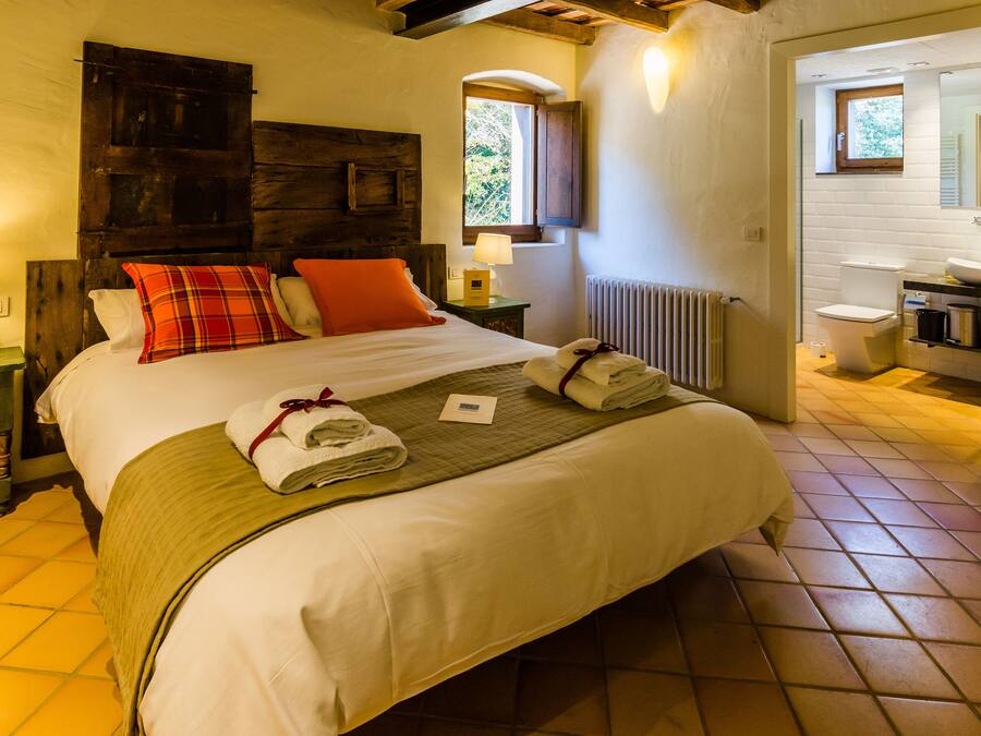 Un dormitorio con una cama grande, cabecera de madera, ventana, mesita de noche, lámpara y vistas al baño.