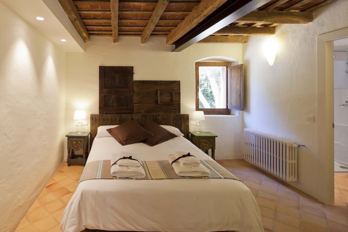 A bedroom with a wooden headboard, two bedside tables with lamps, a radiator, and a window with a view of greenery.