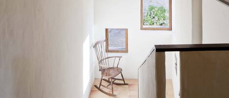 Una escalera conduce a una habitación con una mecedora de madera, dos cuadros enmarcados y vistas al exterior con vegetación.