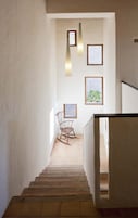 Una escalera conduce a una habitación con una mecedora de madera, dos cuadros enmarcados y vistas al exterior con vegetación.
